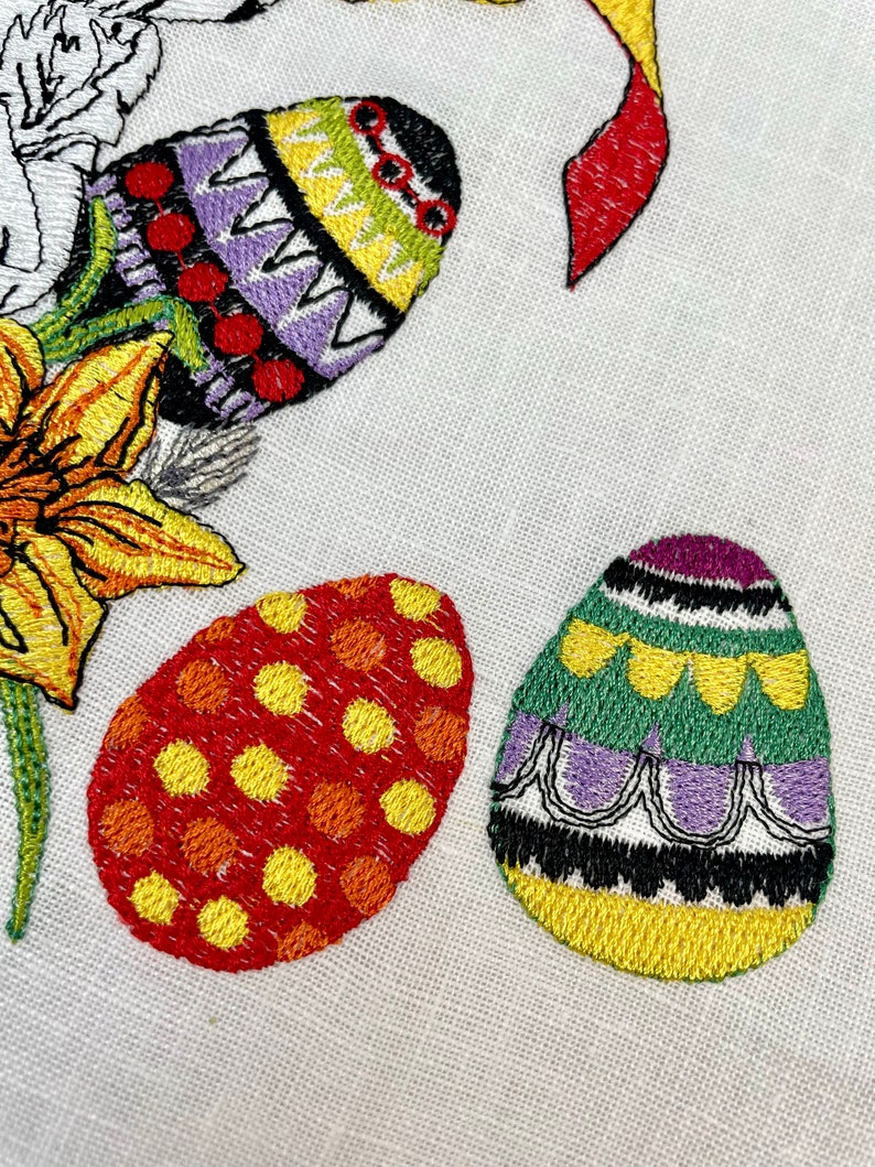 May include: Three embroidered Easter eggs with different patterns. One egg is purple, green, and yellow with a black and white border. Another egg is red and yellow with a polka dot pattern. The third egg is purple, yellow, and green with a zig-zag pattern.