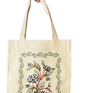 May include: A beige canvas tote bag with a green leafy border and a red and green floral design in the centre. The design features black berries and green leaves.