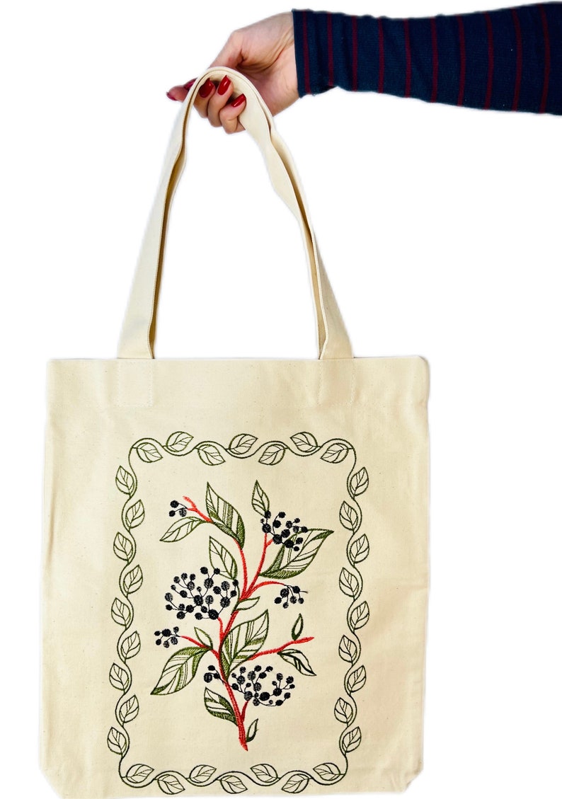 May include: A beige canvas tote bag with a green leafy border and a red and green floral design in the centre. The design features black berries and green leaves.