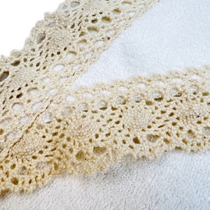 May include: A white towel with a beige crocheted lace trim. The lace trim is detailed with a delicate pattern.