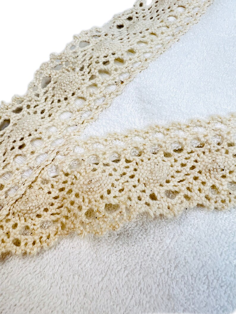 May include: A white towel with a beige crocheted lace trim. The lace trim is detailed with a delicate pattern.