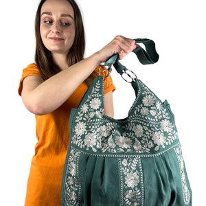 K&ouml;nnte beinhalten: Eine tealfarbene Tragetasche mit wei&szlig;er Blumenstickerei. Die Tasche hat ein plissiertes Design und zwei silberne Metallringgriffe. Eine Person in einem orangefarbenen Oberteil h&auml;lt die Tasche.