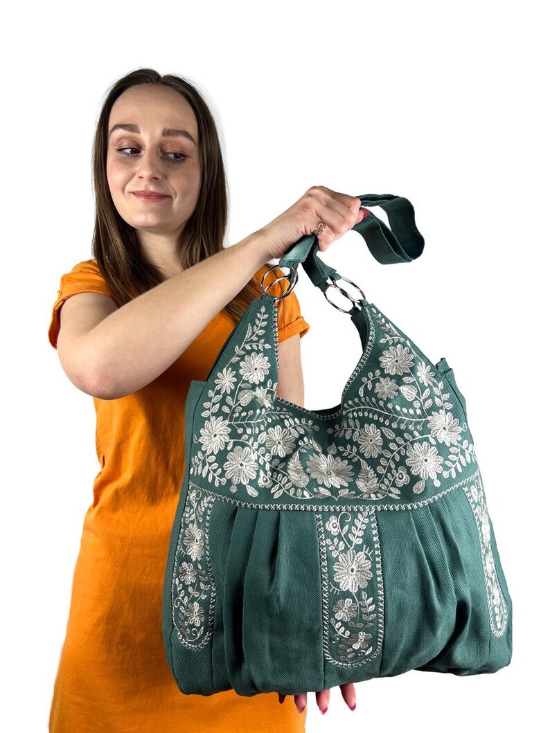 K&ouml;nnte beinhalten: Eine tealfarbene Tragetasche mit wei&szlig;er Blumenstickerei. Die Tasche hat ein plissiertes Design und zwei silberne Metallringgriffe. Eine Person in einem orangefarbenen Oberteil h&auml;lt die Tasche.