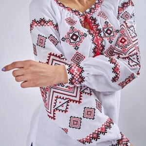 May include: White embroidered blouse with red and black geometric patterns. The blouse has a round neckline and long sleeves.