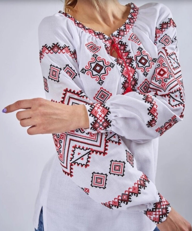 May include: White embroidered blouse with red and black geometric patterns. The blouse has a round neckline and long sleeves.