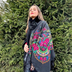 May include: A black shawl with a floral pattern and fringe. The shawl is worn over a black coat and black turtleneck.