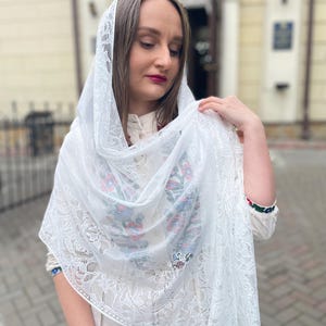 May include: A white lace shawl with floral embroidery in blue, red, and green. The shawl is draped over the shoulders. The edges of the shawl are finished with a delicate fringe.