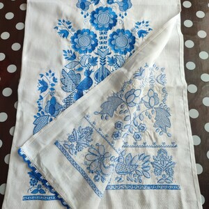 May include: White linen table runner with intricate blue floral embroidery. The design includes stylized flowers, leaves, and a bird. The edges are finished with a blue crocheted trim, adding a delicate touch to the handcrafted textile.