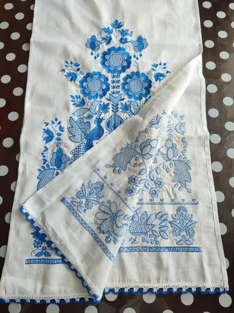 May include: White linen table runner with intricate blue floral embroidery. The design includes stylized flowers, leaves, and a bird. The edges are finished with a blue crocheted trim, adding a delicate touch to the handcrafted textile.