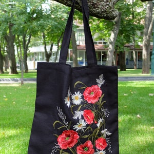 Op de afbeelding: Zwarte canvas tote bag met geborduurde rode klaprozen en witte madeliefjes. De tas heeft een lange band en hangt aan een tak.