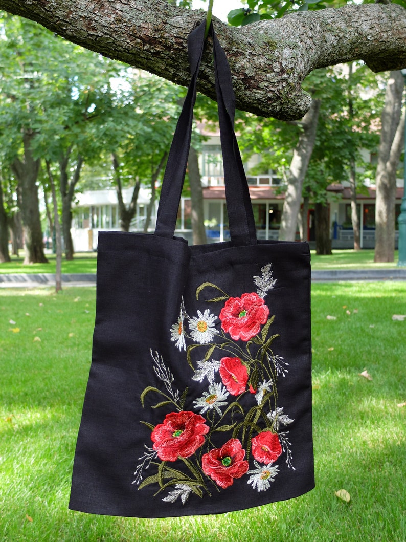 Op de afbeelding: Zwarte canvas tote bag met geborduurde rode klaprozen en witte madeliefjes. De tas heeft een lange band en hangt aan een tak.