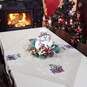 Op de afbeelding: Een beige tafelkleed met geborduurde wintertaferelen en sneeuwvlokken. Een sneeuwpopfiguur met een kleine hoed en een krans van groen, rode bessen en ornamenten staat op de tafel. Een kerstboom en een open haard zijn op de achtergrond.