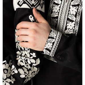 May include: Black shirt with white floral embroidery on the sleeves and collar. The cuffs and upper sleeves feature detailed geometric and floral patterns. The shirt's design includes a tassel detail at the neckline, showcasing traditional craftsmanship.