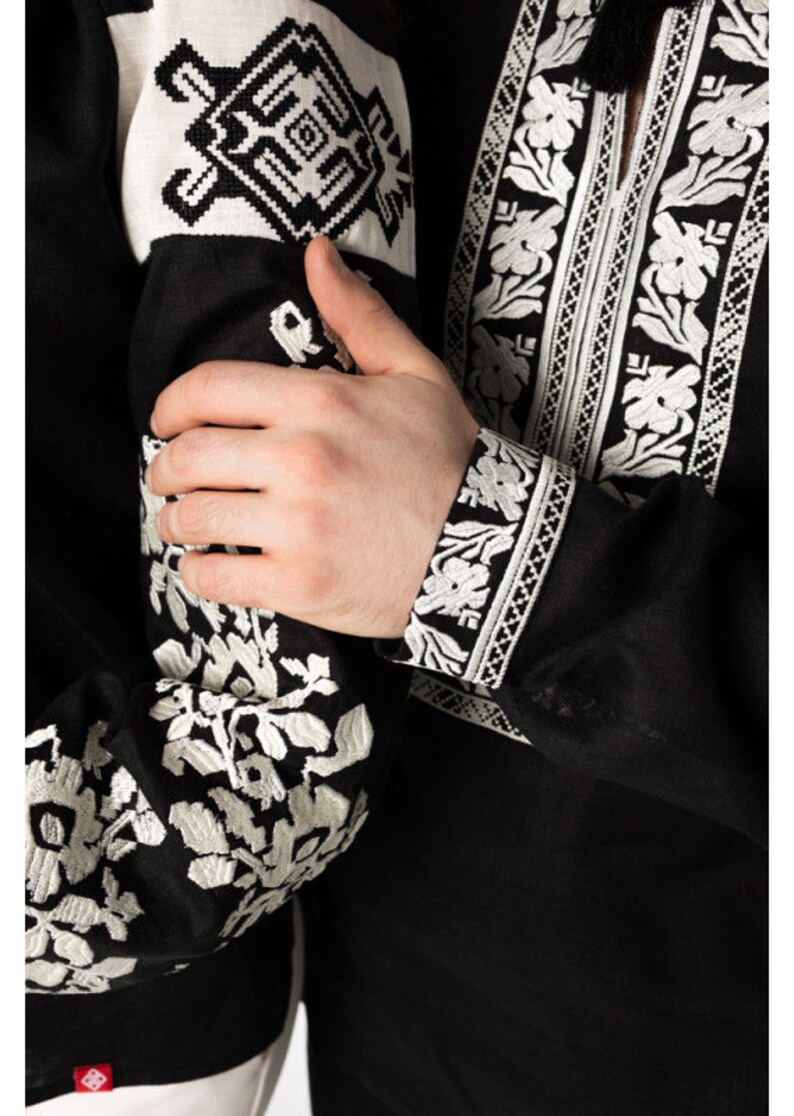 May include: Black shirt with white floral embroidery on the sleeves and collar. The cuffs and upper sleeves feature detailed geometric and floral patterns. The shirt's design includes a tassel detail at the neckline, showcasing traditional craftsmanship.