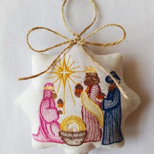 May include: Embroidered star-shaped Christmas ornament featuring the three wise men, a star, and a manger. The ornament is made of white fabric and has a jute twine loop for hanging. The embroidery uses pink, red, blue, yellow, and brown thread.