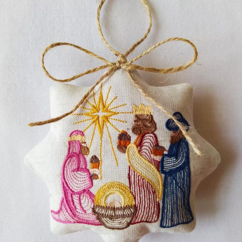 May include: Embroidered star-shaped Christmas ornament featuring the three wise men, a star, and a manger. The ornament is made of white fabric and has a jute twine loop for hanging. The embroidery uses pink, red, blue, yellow, and brown thread.