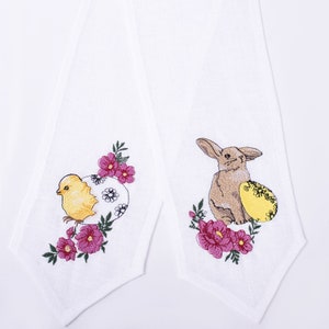 May include: Two white Easter hand towels with embroidered designs. One shows a yellow chick emerging from a floral egg, the other a brown bunny with a yellow egg, surrounded by pink flowers and green leaves.
