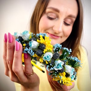 Pode incluir: Uma faixa de cabe&ccedil;a floral com flores azuis e amarelas, segurada nas m&atilde;os de uma pessoa. A faixa de cabe&ccedil;a &eacute; feita de flores e vegeta&ccedil;&atilde;o artificiais.