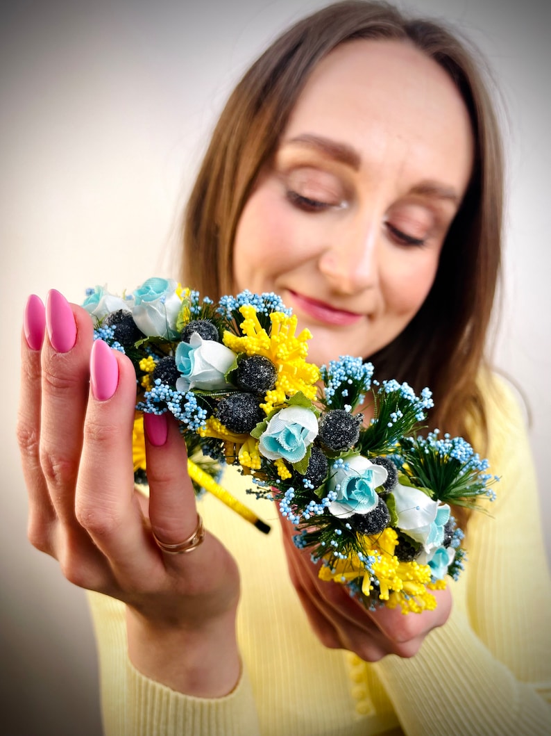 Pode incluir: Uma faixa de cabe&ccedil;a floral com flores azuis e amarelas, segurada nas m&atilde;os de uma pessoa. A faixa de cabe&ccedil;a &eacute; feita de flores e vegeta&ccedil;&atilde;o artificiais.