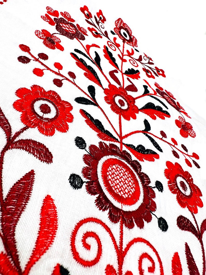 K&ouml;nnte beinhalten: Rote und schwarze Stickerei mit floralem Muster auf wei&szlig;em Stoffhintergrund. Das Design zeigt stilisierte Blumen mit runden Zentren und filigranen Details.