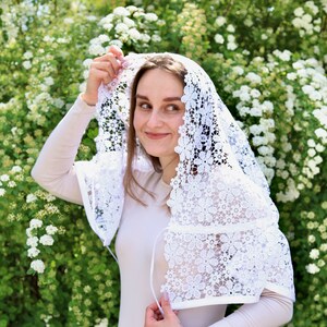 May include: A white lace veil with a floral pattern. The veil is worn over a person's head and shoulders.