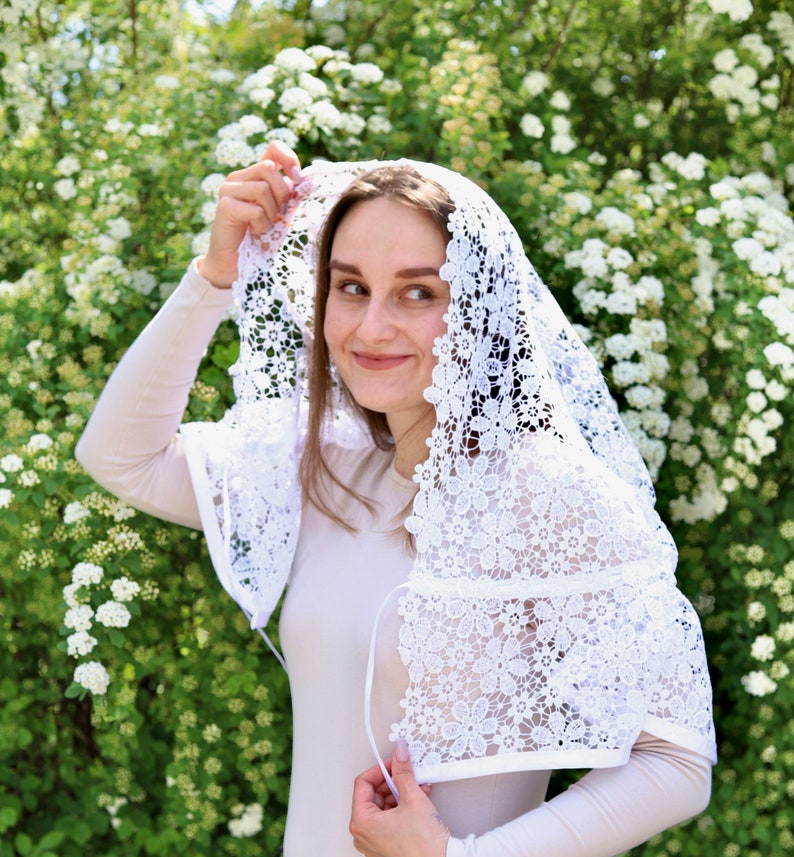 May include: A white lace veil with a floral pattern. The veil is worn over a person's head and shoulders.
