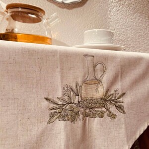 May include: A white tablecloth with an embroidered design of a pitcher and olives. The tablecloth is on a table with a glass teapot and a white cup and saucer.
