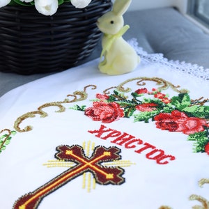 K&ouml;nnte beinhalten: Wei&szlig;es besticktes Tuch mit Kreuz- und Blumendesign, mit dem Wort "Christos" in Rot. Ein schwarzer Weidenkorb mit wei&szlig;en Tulpen und eine gelbe Hasenfigur befinden sich im Hintergrund.