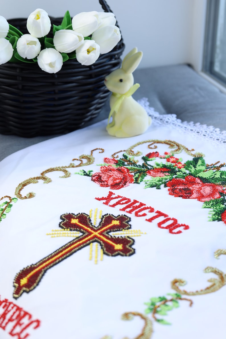 K&ouml;nnte beinhalten: Wei&szlig;es besticktes Tuch mit Kreuz- und Blumendesign, mit dem Wort "Christos" in Rot. Ein schwarzer Weidenkorb mit wei&szlig;en Tulpen und eine gelbe Hasenfigur befinden sich im Hintergrund.