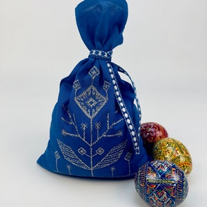 Puede incluir: Una bolsa azul cobalto con cord&oacute;n, bordado floral plateado y ribete blanco. Tres huevos de Pascua decorados en rojo, amarillo y azul est&aacute;n junto a la bolsa. Los huevos tienen patrones intrincados.