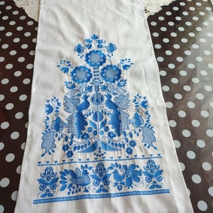 May include: White rectangular linen table runner with blue floral embroidery. The design features stylized flowers, birds, and foliage. The bottom edge has a delicate blue crocheted trim. A traditional, decorative textile.