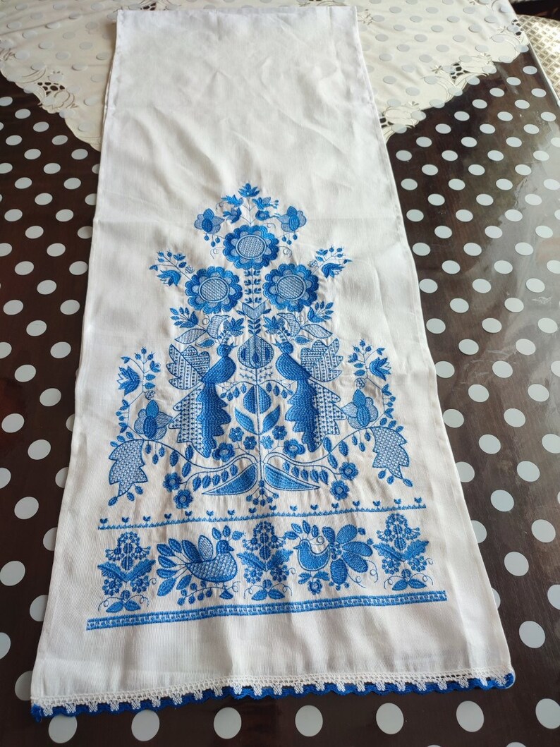 May include: White rectangular linen table runner with blue floral embroidery. The design features stylized flowers, birds, and foliage. The bottom edge has a delicate blue crocheted trim. A traditional, decorative textile.