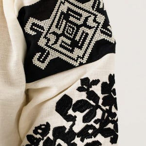 May include: Close-up of a cream-coloured garment with black embroidered details. The design features geometric patterns and floral motifs on the sleeves. The fabric has a textured appearance.