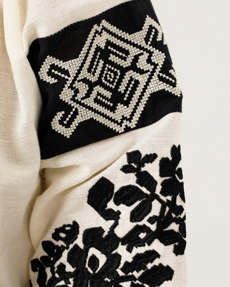 May include: Close-up of a cream-coloured garment with black embroidered details. The design features geometric patterns and floral motifs on the sleeves. The fabric has a textured appearance.