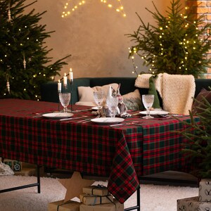 May include: A festive Christmas table setting with a red and green plaid tablecloth. The table is set with plates, glasses, and candles. Presents wrapped in brown paper are stacked on the floor. Christmas trees and a star-shaped light fixture add to the holiday ambiance.