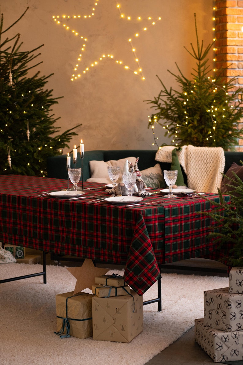 May include: A festive Christmas table setting with a red and green plaid tablecloth. The table is set with plates, glasses, and candles. Presents wrapped in brown paper are stacked on the floor. Christmas trees and a star-shaped light fixture add to the holiday ambiance.