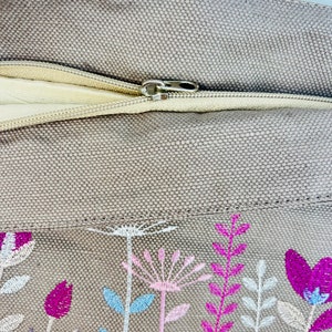 K&ouml;nnte beinhalten: Ein beigefarbenes Kissen aus Canvas mit Rei&szlig;verschluss und gesticktem Blumendesign in Rosa-, Wei&szlig;- und Blaut&ouml;nen.