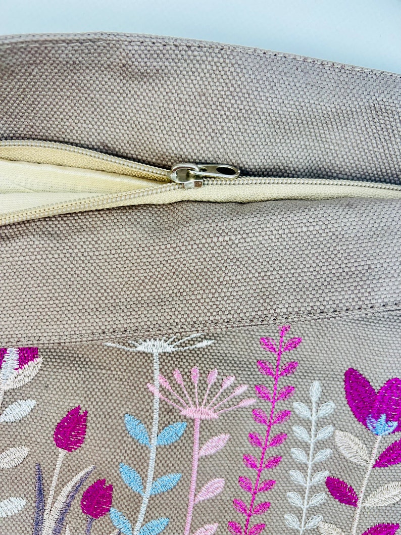K&ouml;nnte beinhalten: Ein beigefarbenes Kissen aus Canvas mit Rei&szlig;verschluss und gesticktem Blumendesign in Rosa-, Wei&szlig;- und Blaut&ouml;nen.