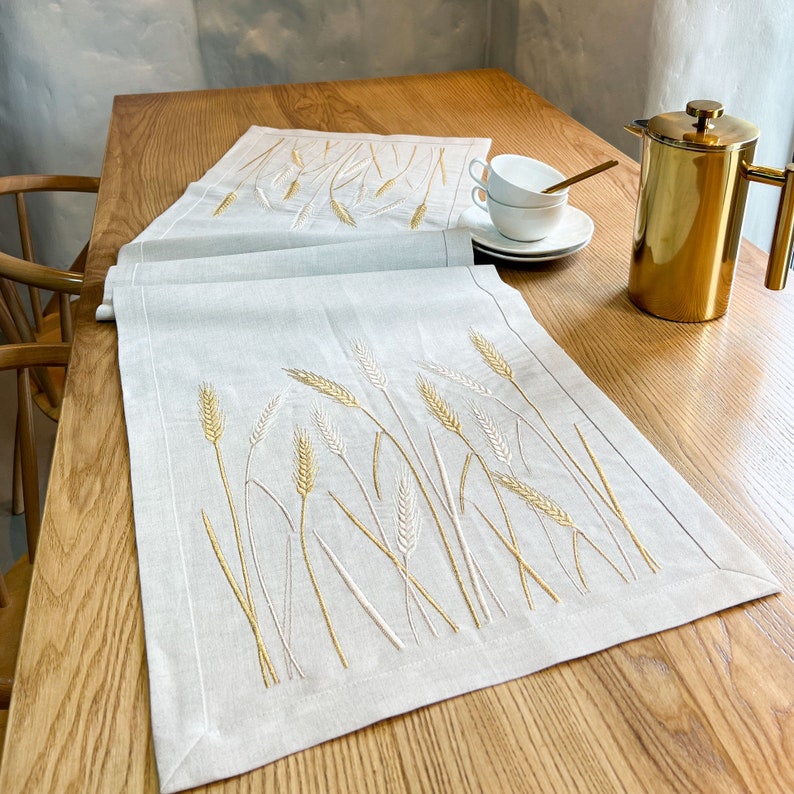 Peut inclure: Deux chemins de table en lin gris clair avec des &eacute;pis de bl&eacute; brod&eacute;s en or et blanc. Les chemins de table sont pos&eacute;s sur une table en bois, avec une cafeti&egrave;re dor&eacute;e et des tasses &agrave; th&eacute; blanches en arri&egrave;re-plan. Les chemins sont rectangulaires.