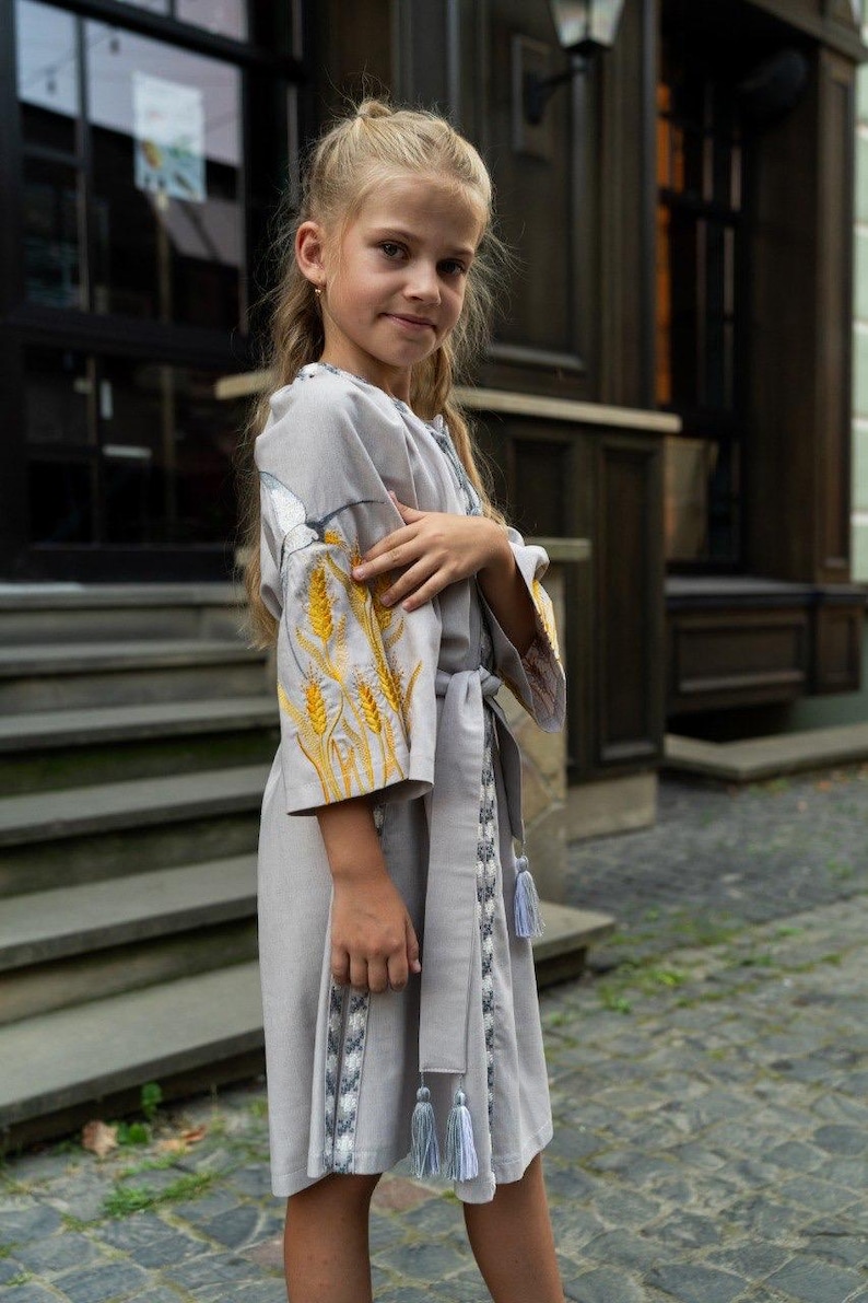 K&ouml;nnte beinhalten: Hellgraues Kleid mit gestickten Weizen&auml;hren auf den &Auml;rmeln. Das Kleid hat einen passenden G&uuml;rtel und dekorative Quasten. Das M&auml;dchen tr&auml;gt das Kleid drau&szlig;en vor einem Geb&auml;ude.