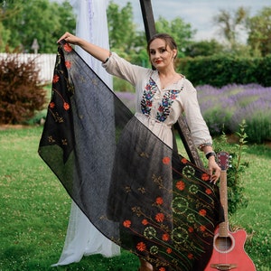 May include: A woman wearing a cream-colored embroidered blouse and a black skirt with floral accents. She is holding a black shawl with orange and green floral designs. A red acoustic guitar leans against a wooden structure.