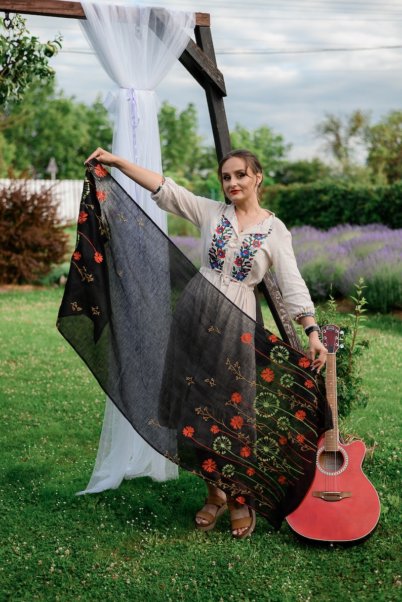 May include: A woman wearing a cream-colored embroidered blouse and a black skirt with floral accents. She is holding a black shawl with orange and green floral designs. A red acoustic guitar leans against a wooden structure.