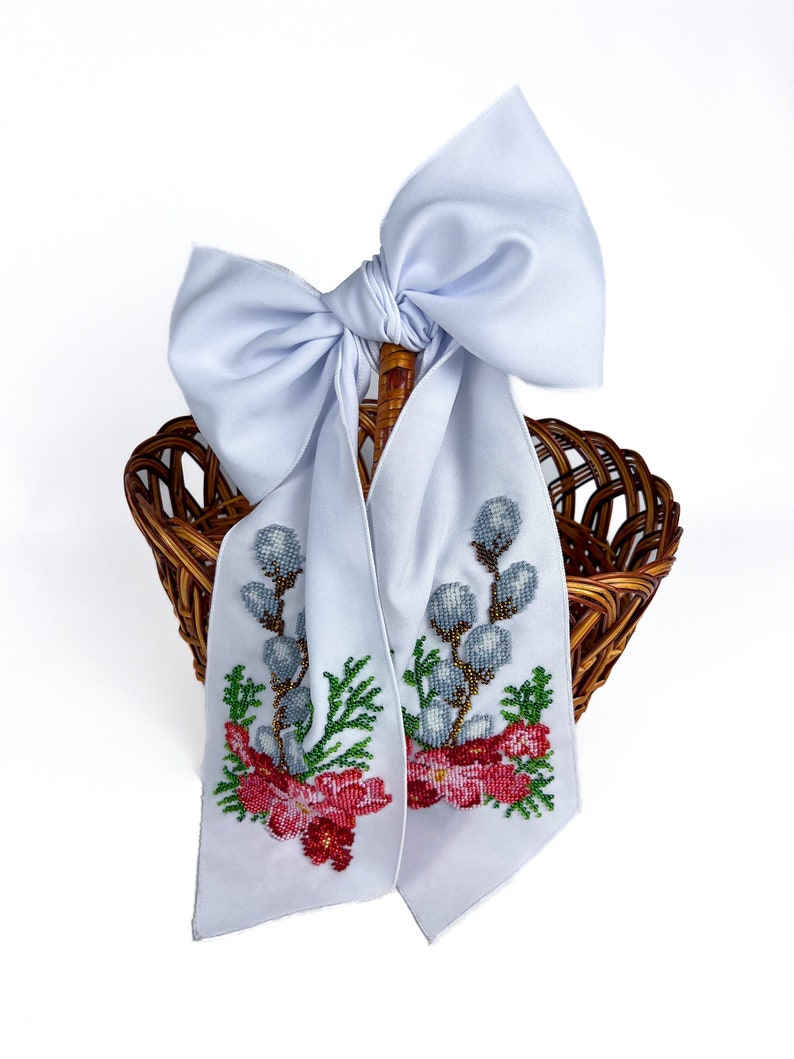 May include: A white wicker basket with a white satin bow and beaded floral embroidery. The embroidery features pink flowers, green leaves, and grey willow branches.