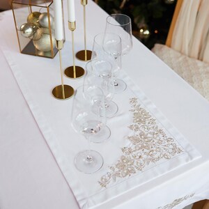 May include: A white tablecloth with a gold embroidered floral design and a matching runner. The table is set with wine glasses, gold candle holders with white candles, and a decorative glass box with gold ornaments.