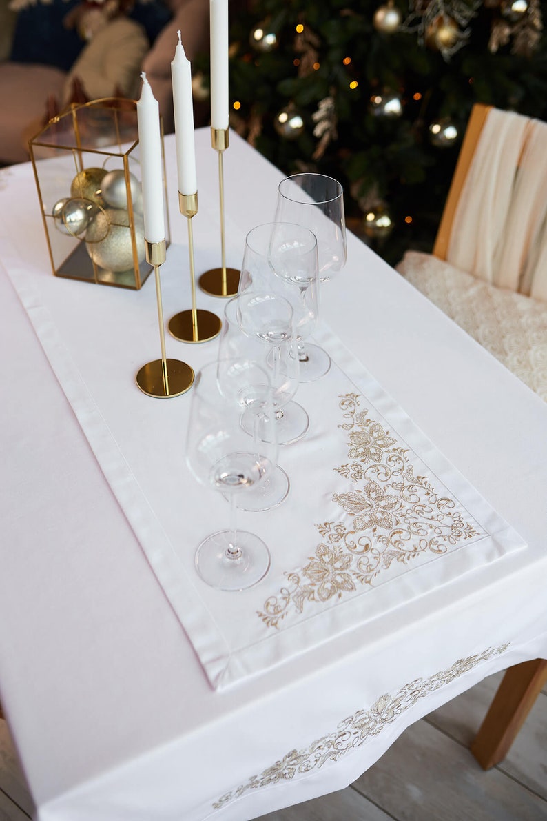 May include: A white tablecloth with a gold embroidered floral design and a matching runner. The table is set with wine glasses, gold candle holders with white candles, and a decorative glass box with gold ornaments.