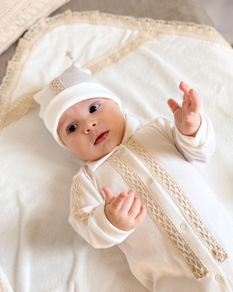 Op de afbeelding: Een witte baby romper met beige kantversiering en een witte gehaakte muts. De romper heeft een knoopsluiting en de muts heeft een klein geborduurd kruis op de bovenkant.