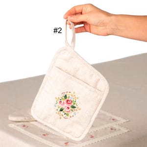 May include: A beige pot holder with a floral design featuring pink roses, green leaves, and blue accents. The pot holder has a pocket and a loop for hanging. The number "#2" is printed on the top left.