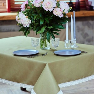 May include: A dining table set for a meal, featuring a light green tablecloth with white lace trim. Two blue plates, silverware, and crystal glasses are set. A vase of pink peonies and candles complete the elegant arrangement.