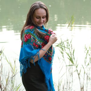 May include: A woman wearing a vibrant blue shawl with a floral pattern of red roses and green leaves. The shawl has a fringed edge and is draped over the shoulders. The background features a lake and tall grass.