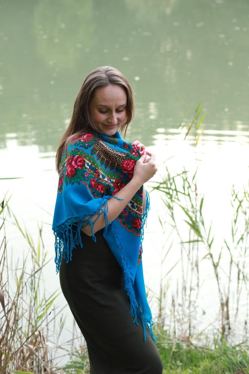 May include: A woman wearing a vibrant blue shawl with a floral pattern of red roses and green leaves. The shawl has a fringed edge and is draped over the shoulders. The background features a lake and tall grass.
